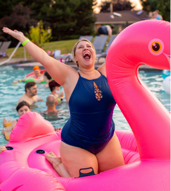 Woman in navy swimsuit looking ecstatic kneeling on blow up pink flamingo in pool